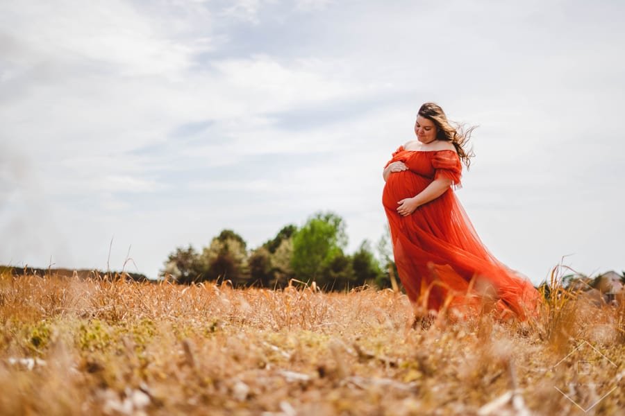 An expectant mother in a flowing red dress standing in a sunlit field